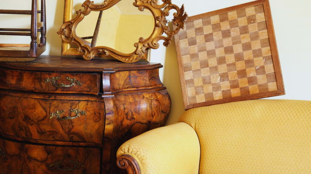Cômoda antiga em madeira entalhada, espelho com moldura dourada, tabuleiro de xadrez de madeira e sofá amarelo, representando móveis usados prontos para venda.
