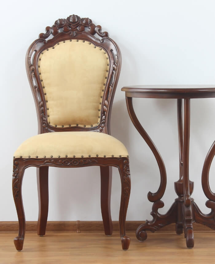 Cadeira e mesa redonda de madeira entalhada em estilo antigo, com assento estofado em tecido bege.